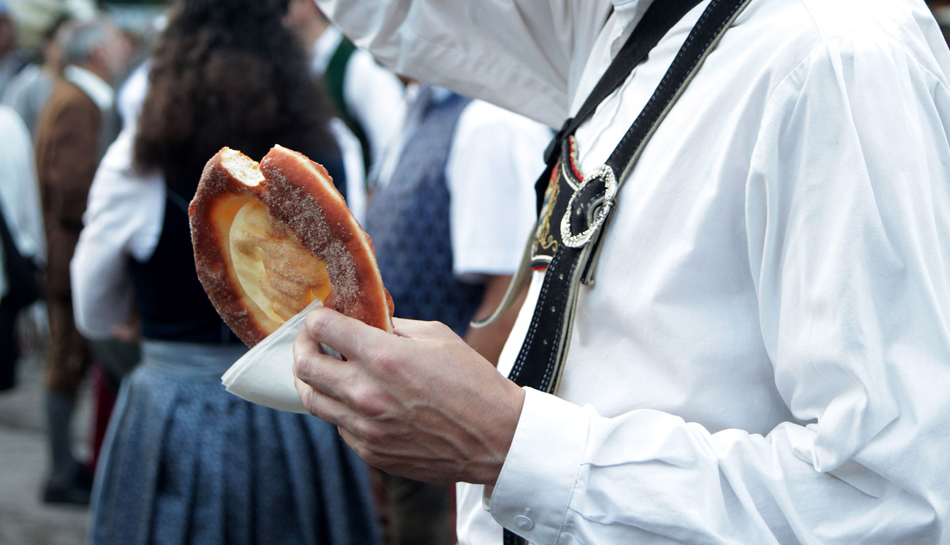Ausschnitt Person in Lederhose und weißem Hemd hält ein Gebäck mit einer Serviette in der Hand