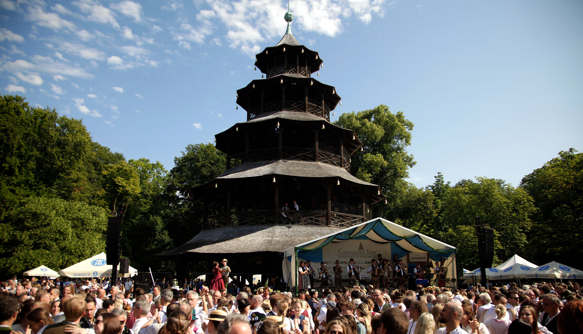 Chinesischer Turm, davor eine überdachte Bühne mit einer Musikkapelle und davor eine tanzende Menschenmenge